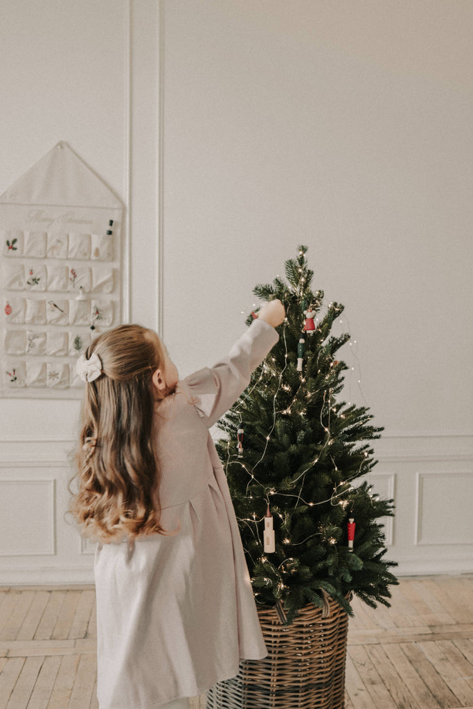 A young girl with long brown hair in a light dress decorates a small Christmas tree with Sabo Concept Christmas Ornaments and lights in a cozy, softly lit room with light walls and wooden floors.