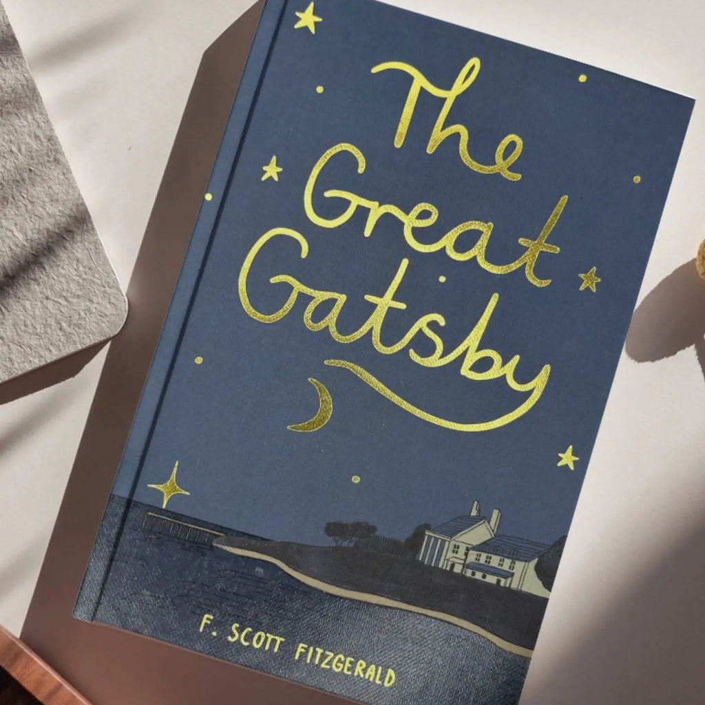 A blue and yellow illustrated hardcover of The Great Gatsby | Collector's Edition rests on a white surface, featuring handwritten-style gold lettering, stars, a crescent moon, and a house by the water.