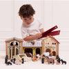 A young child in a white shirt enjoys small world play, arranging animal and building pieces from the Wooden Farmyard Stables set on the floor in front of a white wall.