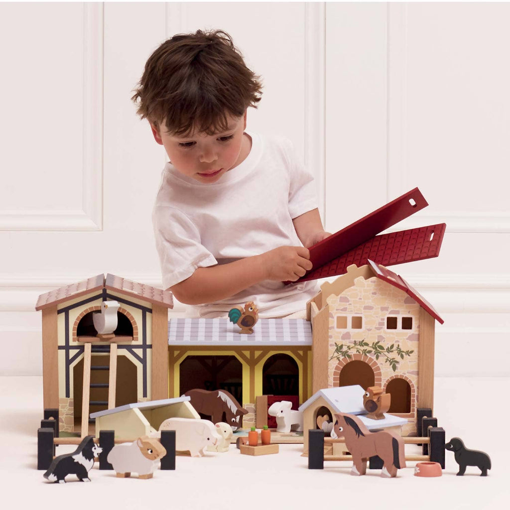 A young child in a white shirt enjoys small world play, arranging animal and building pieces from the Wooden Farmyard Stables set on the floor in front of a white wall.