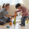 A young girl and a toddler play with the colorful, non-toxic Grimm's Tree Slices Building Blocks on a hardwood floor. The girl, wearing a striped shirt and polka dot leggings, helps the toddler, who is dressed in a striped long-sleeve shirt and jeans, stack the building blocks. Bright, natural light fills the room.
