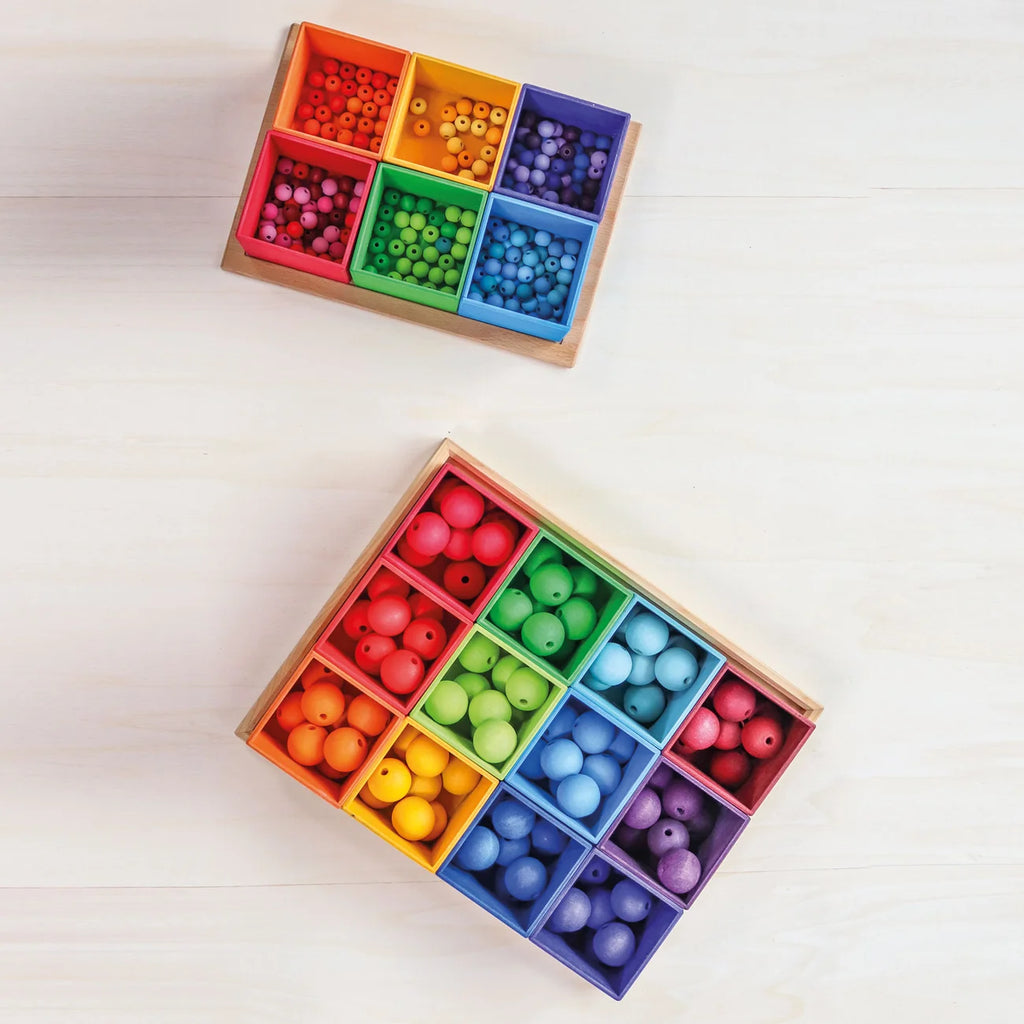 Two natural wood Grimm’s Small 6 Piece Rainbow Box Sorter trays filled with small, colorful beads organized by color into individual square compartments.