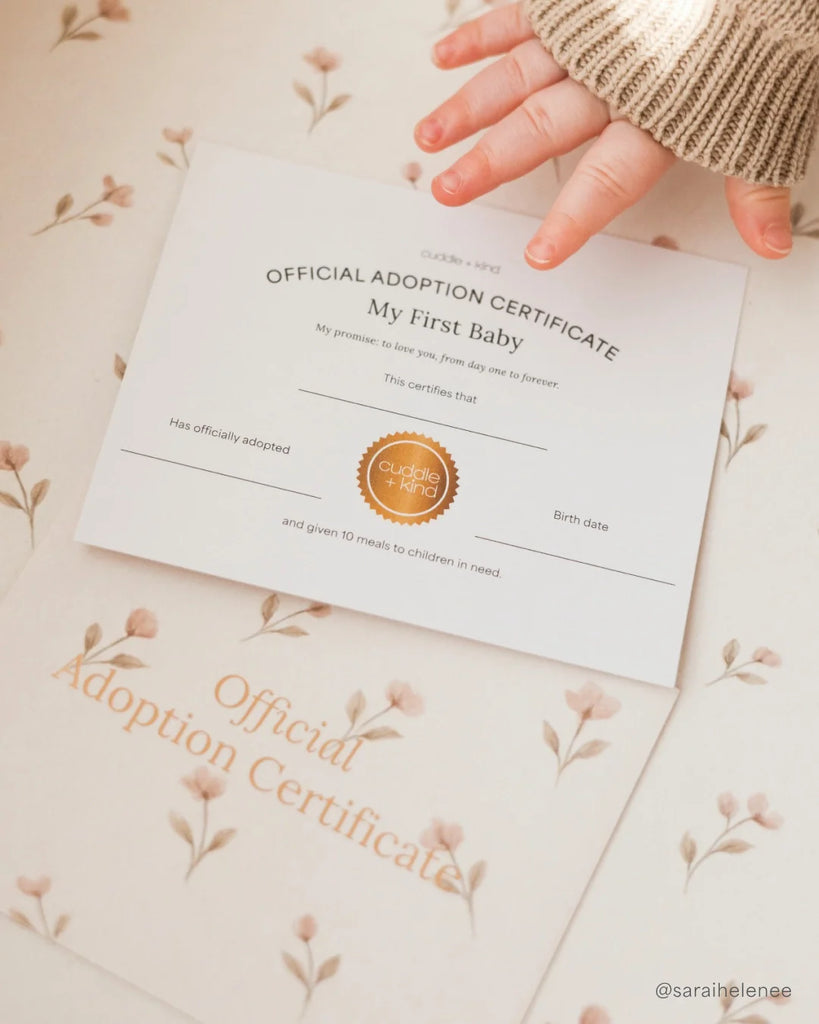 A baby's hand touches an adoption certificate on a floral surface, with another one beneath. Next to them is Cuddle + Kind My First Baby (Buttercup), a hand-knit natural cotton doll that donates 10 meals to children in need. Ships in one week.