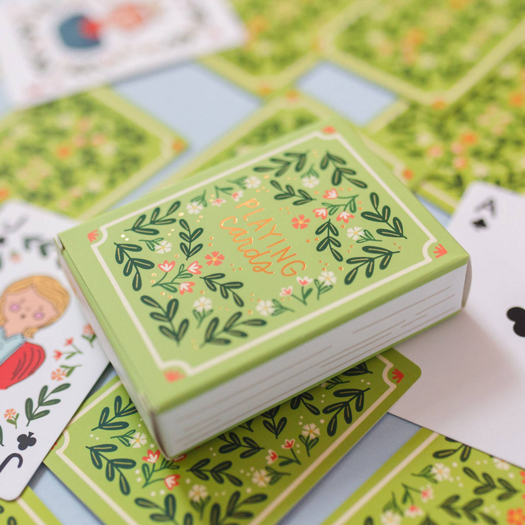 A green box of the Green Floral Deck of Playing Cards (ships in approximately one week) rests on scattered cards, some face up to display hand-illustrated floral designs and classic suits—a delightful choice for card game lovers.