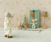 A small, plush rhinoceros in pajamas stands on a carpet, facing a plush mouse from the Maileg mice family dressed in striped clothes. The mouse is near the Maileg Mouse Closet in soft blue, adorned with golden hangers and topped with a toy carrot. The room features flowery wallpaper and a small floor lamp.