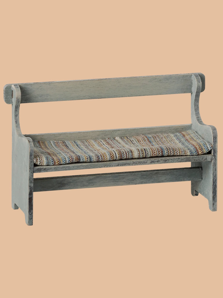 A small wooden bench with a simple backrest and colorful woven seat from the Maileg Garden Hideaway Set sits beside a Mouse Size Maileg Flower Pot With Plant against a plain peach background.
