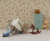 In a playful scene, a charming mouse doll in a striped shirt relaxes on a cushion, surrounded by the Maileg Mouse Rocking Horse, a blue cupboard, yellow chair, clock, and miniature accessories, all set against patterned wallpaper.