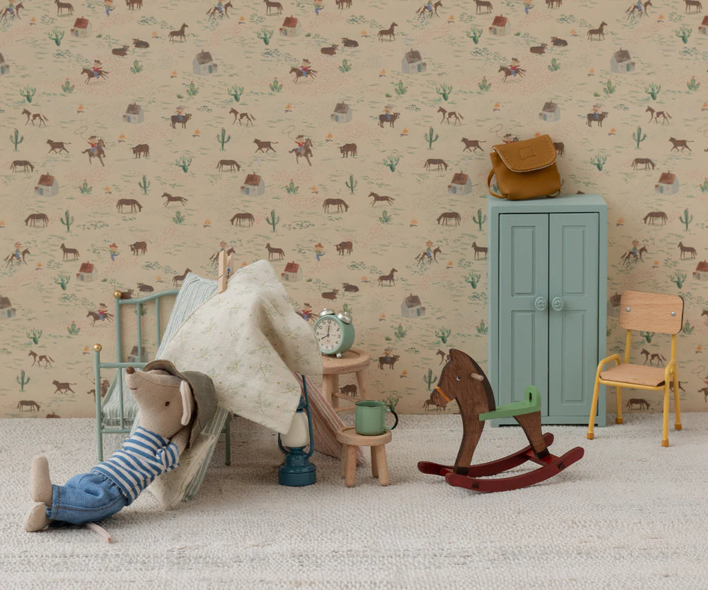 In a playful scene, a charming mouse doll in a striped shirt relaxes on a cushion, surrounded by the Maileg Mouse Rocking Horse, a blue cupboard, yellow chair, clock, and miniature accessories, all set against patterned wallpaper.
