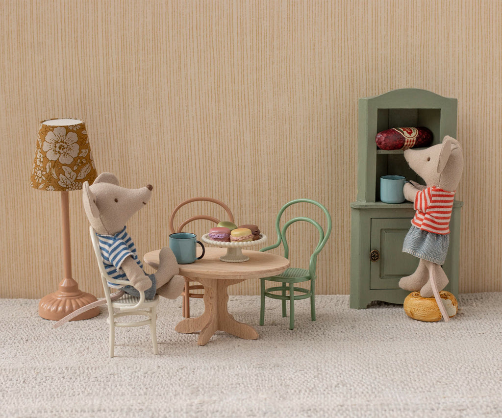 Two toy mice in a cozy dollhouse; one sits on the Maileg Vintage Chair, Mouse Size - Off White by a table with mugs and pastries, while the other stands on a stool reaching into a green cabinet. A vintage lamp is nearby.