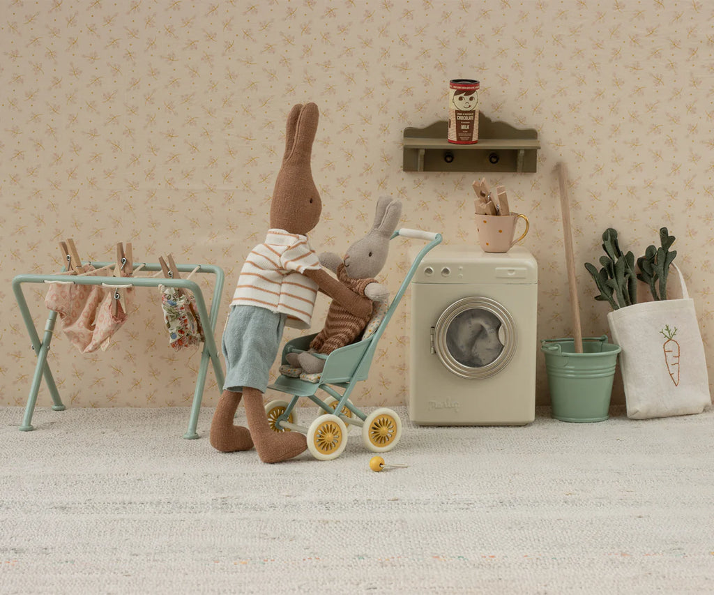A stuffed bunny in a striped shirt and blue shorts pushes a smaller bunny in the Maileg Stroller, Micro - Mint. Mini laundry hangs on a rack, with a toy washing machine and plants adding to the cozy scene.
