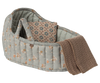 The Maileg Carry Cot, Large - Blue features a soft, quilted design with a blue floral pattern, includes a matching pillow, and comes with a brown knitted blanket draped over the side.