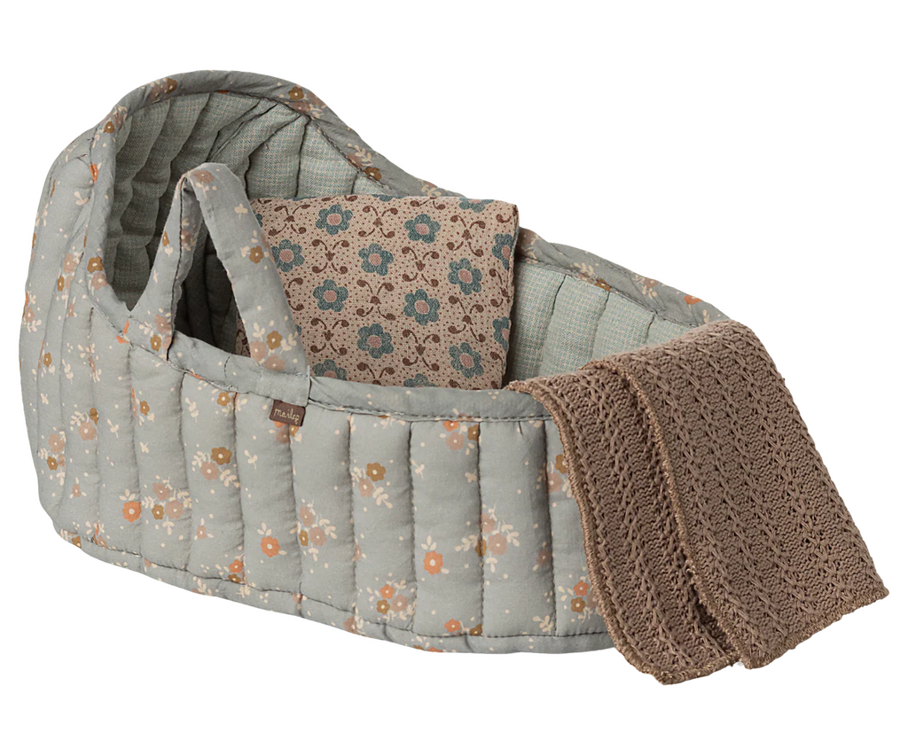 The Maileg Carry Cot, Large - Blue features a soft, quilted design with a blue floral pattern, includes a matching pillow, and comes with a brown knitted blanket draped over the side.