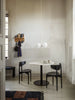 A minimal dining area features a round white table with black base, two black chairs, a modern white pendant lamp, and a Ferm Living Place Rack - Small displaying kitchenware on a light-painted wall. Natural light filters in from a nearby window.