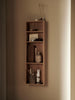 A vertical, wall-mounted Ferm Living Bon Shelf - Oiled Oak with various aesthetic storage solutions including books, a vase with dried branches, and small storage containers, illuminated by soft light from the right.