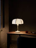 The Poem Table Lamp - White/Cashmere features a translucent shade and pull chain switch, elegantly resting on a wooden table in a dimly lit room. A small red container and closed notebook enhance the Baroque ambiance, while part of a chair is visible in the foreground.