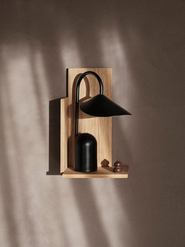 The Ferm Living Arum Portable Lamp in black, featuring a curved neck, sits on a light wooden wall shelf against a brown wall. Next to it is a small brown chess pawn, with the lamp’s soft LED glow casting gentle shadows.