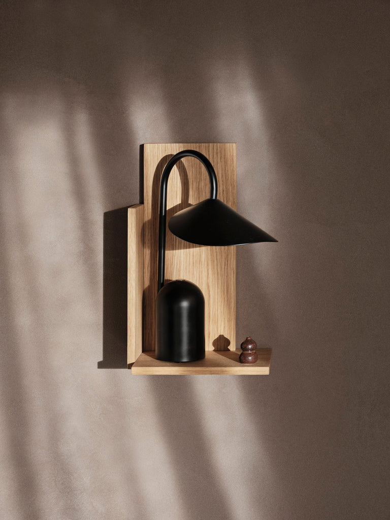 The Ferm Living Arum Portable Lamp in black, featuring a curved neck, sits on a light wooden wall shelf against a brown wall. Next to it is a small brown chess pawn, with the lamp’s soft LED glow casting gentle shadows.