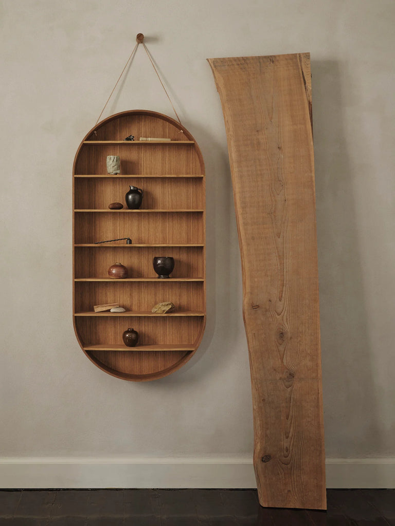 The Ferm Living Oval Oak Dorm Shelf features five minimalist tiers, ideal for displaying vases, stones, or figurines. This tall oak shelf leans against a textured beige wall, adding modern storage and style to any space.