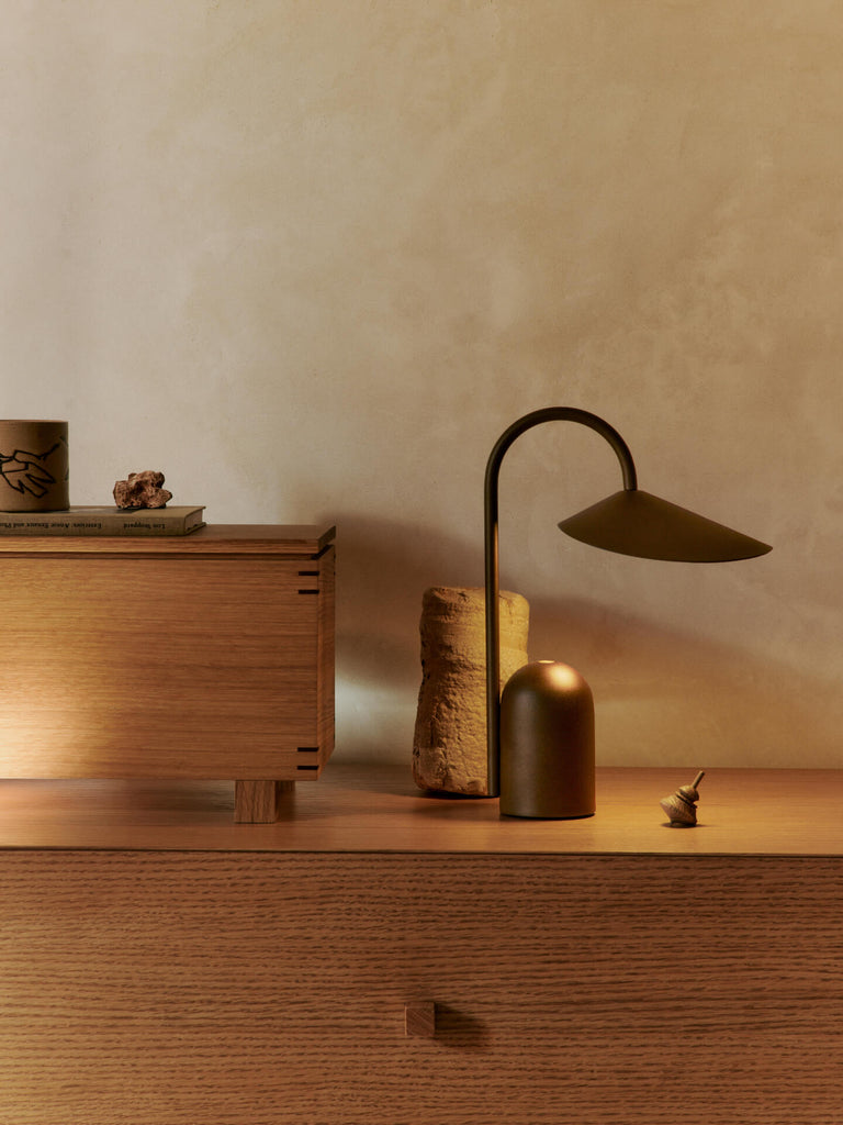 A minimalist wooden sideboard displays the Ferm Living Arum Portable Lamp in Dark Chocolate with touch dimmer, a small cylindrical sculpture, spinning top, and decorative box. Warm lighting adds coziness against a neutral beige wall.