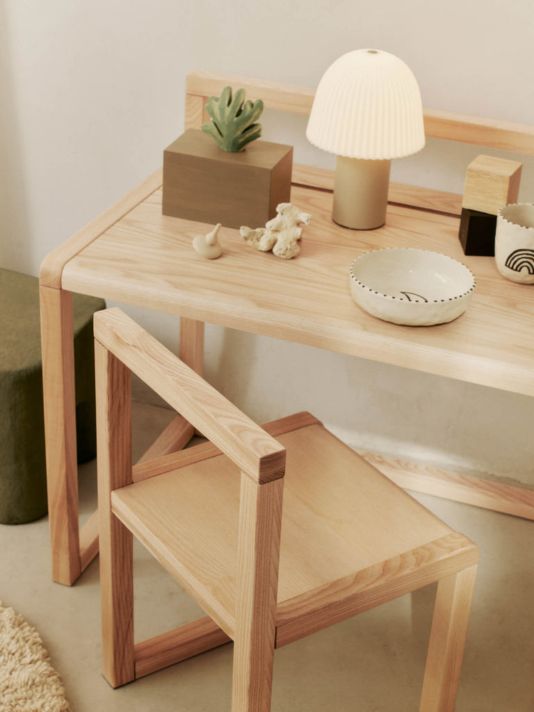 A light wood desk with a matching chair, adorned with the Ferm Living Frill Portable Lamp in Cashmere/White, a ceramic bowl, plant, and abstract decor on a beige floor with an olive green cushion nearby.