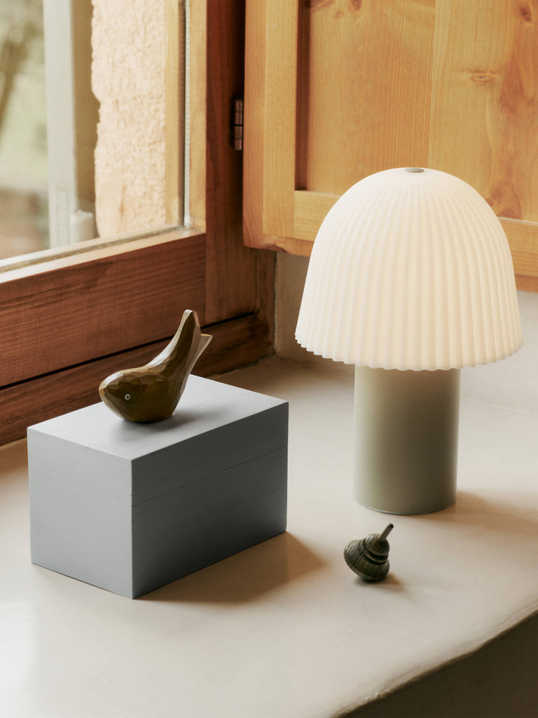 A windowsill with a gray box, small ceramic bird, striped sea shell, and a Ferm Living Frill Portable Lamp in Cashmere/White with a pleated shade—perfect for adding charm to a kids’ room beside the wooden-framed window.