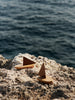The Ferm Living Opty Boat - Set of 2 features small decorative boats made from FSC-certified oak with brown triangular sails, displayed on a rocky shore with the ocean softly blurred in the background.