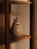 The Ferm Living Pear Money Bank, a ceramic piggy bank with a coin slot, sits on a wooden shelf in warm lighting against vertical textured panels—a charming decor piece for any children's room.