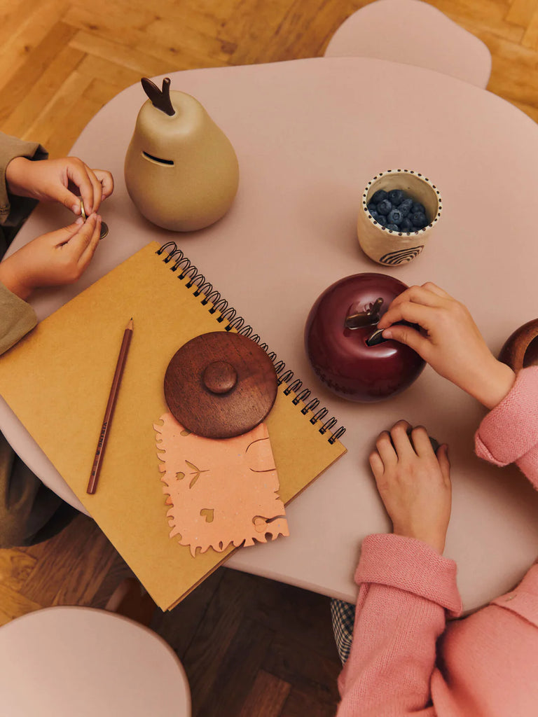 Two children sit at a light pink table with a notebook, pencil, blueberries, cleaning cloth, and playful wooden containers—including the Ferm Living Pear Money Bank—perfect for adding charm to kids' room decor. Both eagerly reach for the containers.