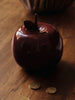 A glossy, dark red Ferm Living Apple Money Bank with a stem top rests on a wooden surface beside gold coins—perfect as a ceramic piggy bank or charming kids’ room decor. Ships in one week.