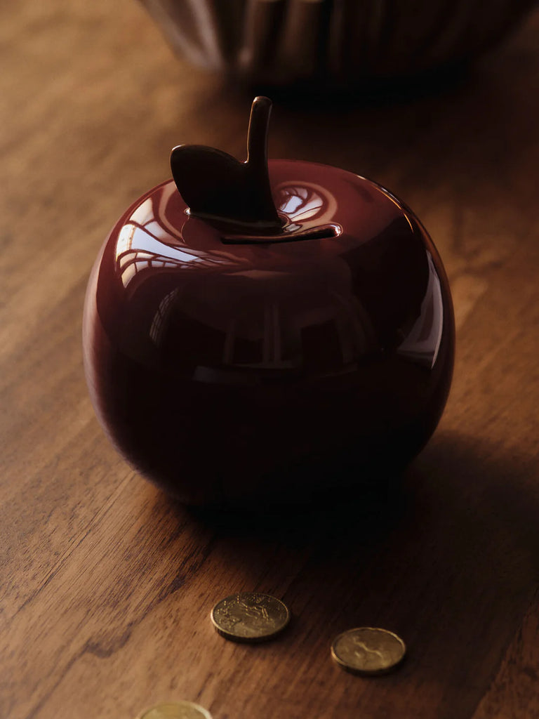 A glossy, dark red Ferm Living Apple Money Bank with a stem top rests on a wooden surface beside gold coins—perfect as a ceramic piggy bank or charming kids’ room decor. Ships in one week.