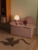 A small mauve storage bench against a green wall holds a ribbed white lamp, the Ferm Living Apple Money Bank (ships in one week), and several wooden toys. A beige rug with a brown abstract pattern covers the wooden floor.