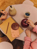 Two children’s hands rest on a small pink table with a spiral notebook, pencil, cup of blueberries, cloth, and the Ferm Living Apple Money Bank—ships in one week—perfect as kids room decor, all neatly arranged on the tabletop.