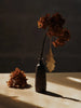 A Ferm Living Oak Leaf Ornaments - Set of 4 - Brass hangs from a dried hydrangea stem in a narrow vase.