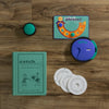 Board Game: Catch Phrase Vintage Bookshelf Edition shown on a wooden surface with a green book-shaped box, circular game unit, buzzer, two white discs, and a green-orange scorecard.