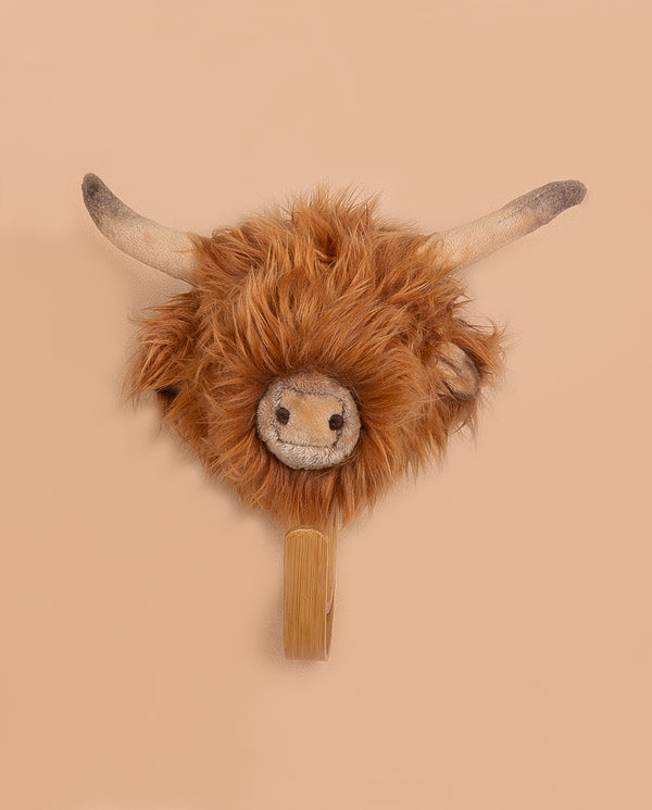 The Coat Hanger - Highland Cow is a plush wall-mounted decoration with long horns, shaggy brown fur, and a smiling face, doubling as an adorable coat hanger against a plain beige background.