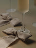 Two natural Ferm Living Bow Decoration - Small - Set of 2 - Natural (Organic Cotton) tied around the stems of two fluted glasses on a table.