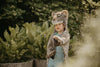 A young child wearing the Costume - Leopard stands outdoors among lush green plants, reaching forward and gazing off to the side.