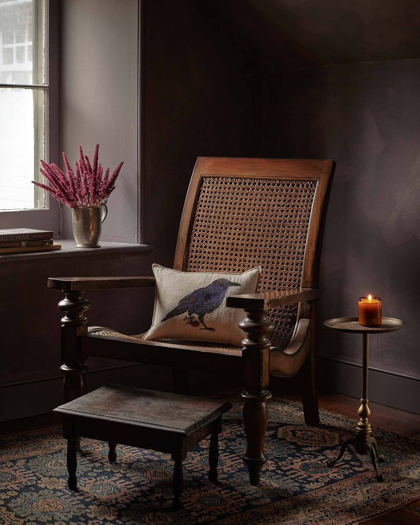 A wooden armchair with a woven back and a Coral & Tusk Blackbird with Acorn Pillow sits by a window with purple flowers, next to a lit candle and footstool on a patterned rug.