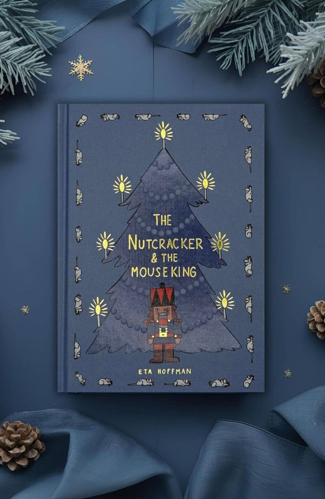 A blue hardcover of "The Nutcracker | Collector's Edition" rests on a blue surface with pinecones and evergreen, featuring the iconic Nutcracker and Christmas tree on the cover.