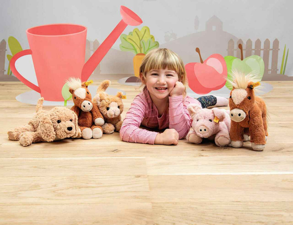 A smiling young girl lies on a wooden floor surrounded by five stuffed animals, including a dog, horse, lion, and the 11-inch Steiff Piko Pig Plush Toy, with a colorful cartoon garden backdrop behind her.