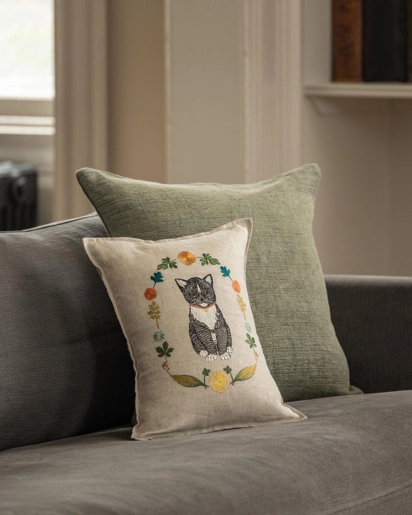 The Coral & Tusk Kitten Cameo Pillow, embroidered with a Tuxedo Cat and colorful flowers, rests against a plain green cushion on a gray sofa in a softly lit room.