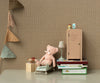 A cozy nursery scene featuring the Maileg Teddy - Rose snuggled in a tiny bed of soft fabric, surrounded by children's books, a small cabinet, and a cupcake. Colorful triangular flags hang against patterned brown wallpaper, adding a playful touch. Note that this product ships in approximately one week.