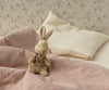 The Maileg My First Bunny - Brown, a soft plush toy with a ribbon, sits on a bed with pink checkered blanket and cream pillows against floral wallpaper—arriving in a beautiful gift box perfect for any special occasion.