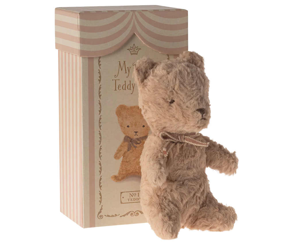 The Maileg My First Teddy, Powder, a small plush bear with a vintage look and brown ribbon, sits beside a striped cardboard box. This perfect newborn gift features an illustration of the teddy bear and decorative text.