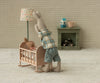 A Maileg Rabbit, Size 1 - Classic - Shirt and Shorts kneels beside a crib, comforting a smaller stuffed rabbit in a cozy, vintage-style room with miniature furniture, a lamp, fireplace, mug, and playful decor.