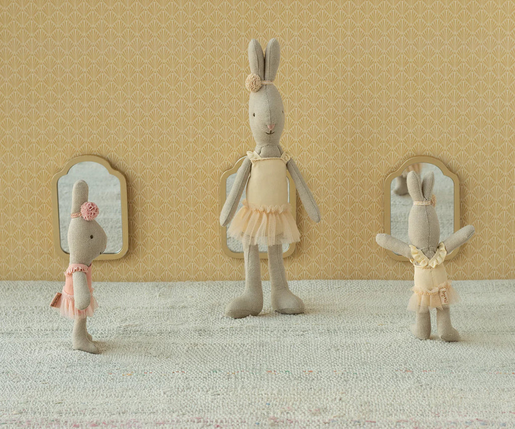 Three Maileg Rabbit, Size 1 - Classic - Ballet Suit and Skirt - Cream toys in pastel tutus stand on a textured surface before a patterned yellow wall with three small mirrors, striking playful poses as if mid-dance.