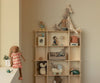 A wooden shelf displays soft toys made from natural fabrics, including rabbits, a penguin, and a giraffe. On the left edge sits the Maileg Bunny, Size 3 - Chocolate Brown - Dress in a softly lit, beige-colored room.