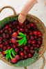 A child’s hand reaches into a woven basket of ripe cherries—some are actually Mery the Cherry Teether baby toys, made from natural rubber with green plastic leaves—resting on green leaves against a light background.