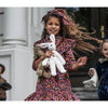 A smiling young girl in a colorful floral dress holds the Senger Naturwelt Stuffed Animal - White Bunny, handmade in Germany from organically grown cotton, while standing outside with other children as her curly hair blows in the wind.