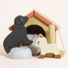 The Dolls House Pet Set features a wooden dog and cat in front of a small doghouse with a red roof, ideal for imaginative play. A blue bowl with a bone design and a soft gray object inside the cozy house complete the set.